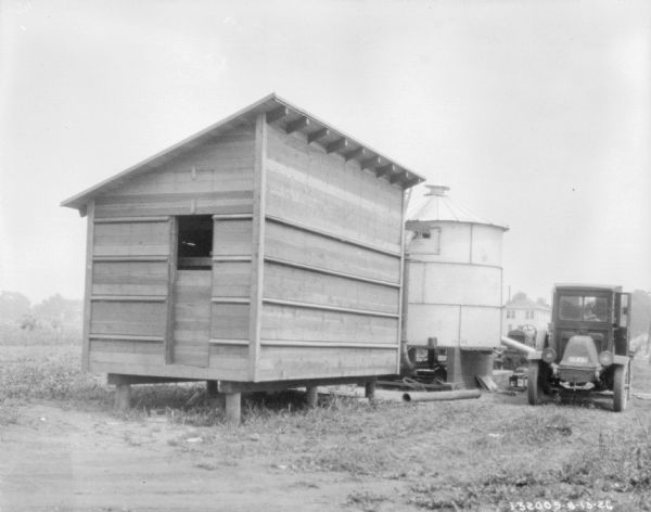 View of a corn crib set on a platform above the ground. Just behind the corn crib is a round, metal storage building with a roof. On the right is a truck, a tractor, and in the far background, a farmhouse.