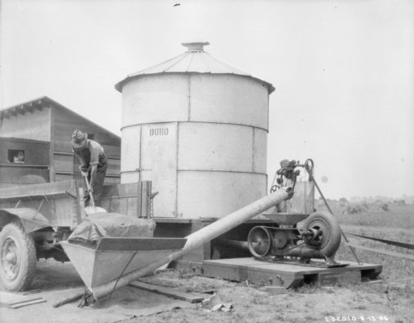 A man is standing in the back of a truck bed shoveling something into a hopper. There is a child looking out of the rear window of the cab. The hopper is attached to a pipe that leads up to belt-driven machinery set up next to round, metal storage silo. A painted sign on the door to the storage silo reads: "Duro." Behind the truck next to the silo is a wooden corn crib.