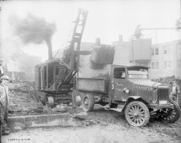Two men are standing near a truck that is being filled by a steam shovel. A sign painted on the side of the front of the truck reads: "Plack Transfer." There is a building in the background on the right.