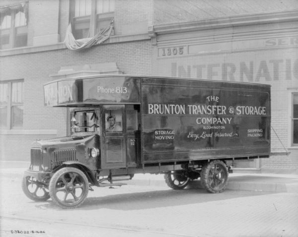 Three-quarter view from front left of a man sitting in the driver's seat of a delivery truck. Signs painted on the side of the building read: "The Brunton Transfer & Storage Company, Bloomington, Every Load Insured." In the background is a large, brick building.