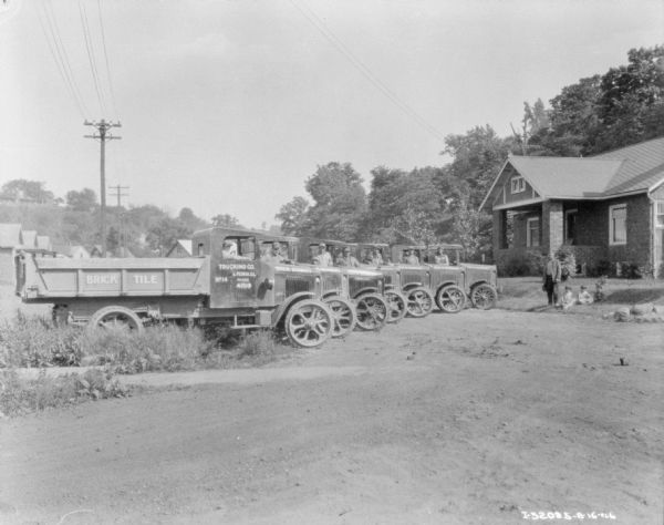 View down road towards men standing with a fleet of trucks parked in a row at an angle. Signs painted on the sides of the truck read: "Brick, Tile," and "Geo. H. Schmidt," and "Trucking Co." A man is standing on the right on a lawn near three children, who are sitting on the ground. Behind the man and three children is a brick building with a porch.