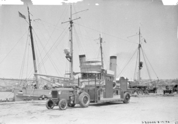 A man is driving an industrial tractor to pull a large trailer along a shipping dock. There is a ship alongside the dock in the background.