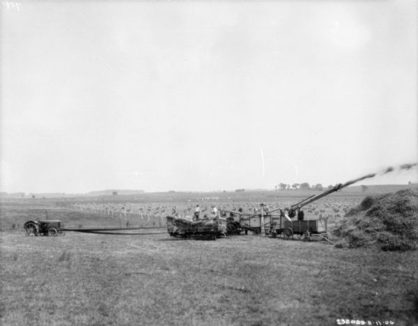 Elevated view across field towards man working with machinery. A tractor is belt-driving the threshing machine.