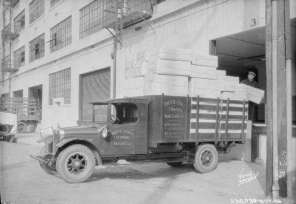 Delivery truck at a loading dock, which is piled high with packages wrapped with paper and string. The sign on the side of the truck reads: "Pacific Coast Terminal Warehouse."