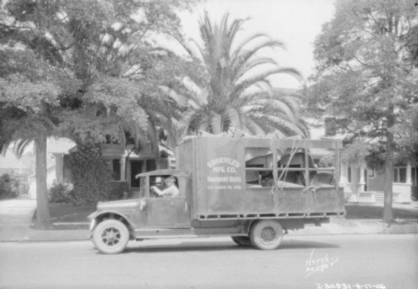 View across street towards a man driving a delivery truck in a suburban neighborhood. with palm trees along the sidewalk. The sign painted on the side of the truck reads: "Kroehler Mfg. Co. Davenport Beds."