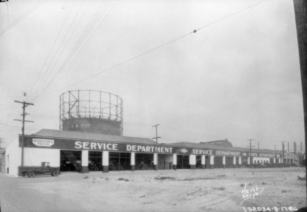View across lot towards the service department for International trucks. There is a truck parked nearby on the left. Behind the building is what may be a large water tower.