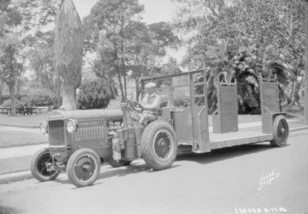 A man is driving a McCormick-Deering industrial tractor to pull a large trailer on a street. In the background is what may be a park, with many types of trees.