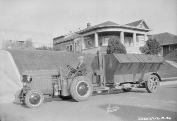 View across street towards a man driving a McCormick-Deering tractor pulling a garbage wagon in a neighborhood. There is a house on a hill in the background.