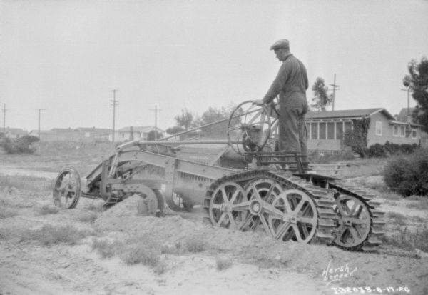 Man driving a grader in a field. There are buildings in the background.