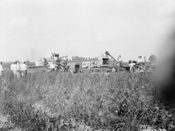 View across field towards men standing near a threshing operation. There are two tractors, each pulling a harvester thresher.
