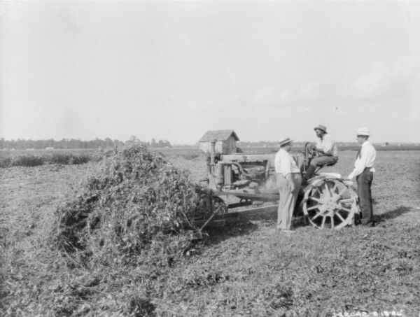 Two men are standing and talking with a man on a tractor.