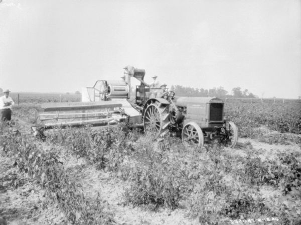 A man is driving a tractor pulling a man on a harvester thresher in a field. Another man is standing on the left watching.