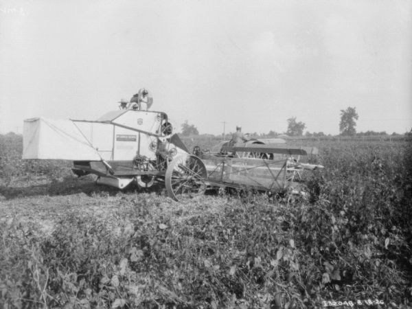 Three-quarter view from right rear of a harvester thresher being pulled by a tractor in a field. There is a man driving the tractor, and another man on the harvester thresher.