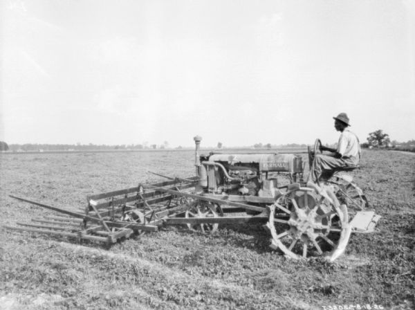 Left side view of a man driving a Farmall tractor in a field with a sweep rake mounted on the front.