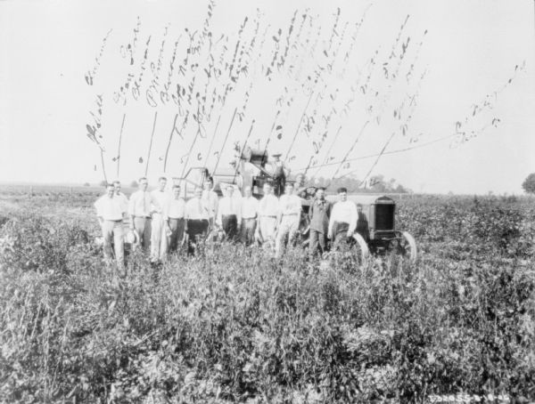 A large group of men are posing in a field with a harvester thresher and a McCormick Deering tractor. The copy of this photograph has writing that numbers and names the men in the group portrait.