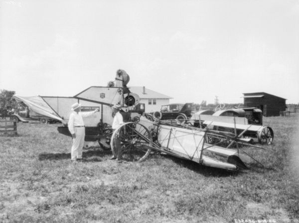 Two men are standing near a harvester thresher attached to a tractor parked in a field. There are automobiles and buildings in the background.