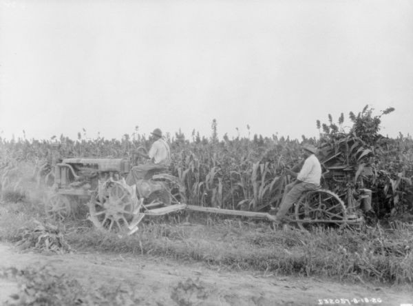 Left side view of a man driving a Farmall tractor that is pulling a man on a corn binder in a field.