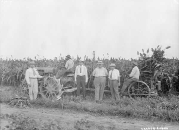 A group of four men are posing in front of a man driving a Farmall tractor pulling a man sitting on a corn binder in a field.