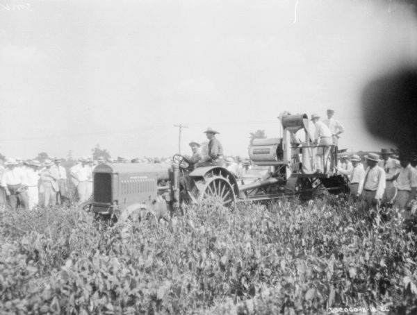 A large group of men are gathered around a man driving a McCormick-Deering tractor pulling a harvester thresher in a field.
