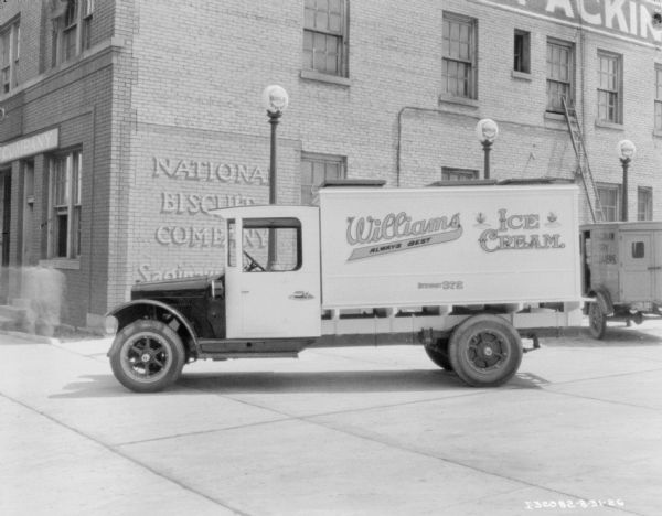 View across street towards a delivery truck for Williams Ice Cream parked near a building. A sign painted on the brick building in the background reads: "National Biscuit Company." There is a sign on a post behind the truck that reads: "Valvoline Motor Oils." A truck parked parked in the background on the right has a sign painted on the side that reads: "Saginaw Dry Cleaners."