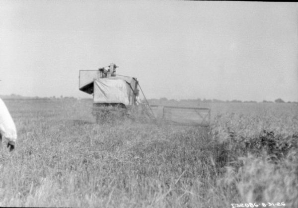 Rear view of a harvester thresher working in a field. There is a person standing on the far left in the foreground.