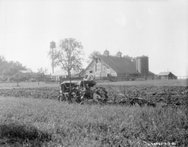 Three-quarter view from left rear of a man driving a Farmall tractor pulling a plow in a field. In the background are farm buildings and a water tower.