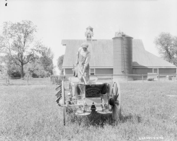 Rear view of a man standing on a lime spreader attached to a Farmall tractor. In the background is a silo and a barn.