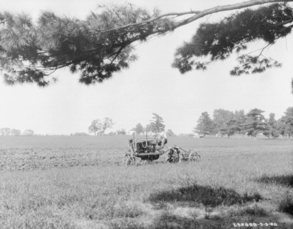 View across field towards a man driving a Farmall tractor pulling a plow. Farm buildings are in the far background.