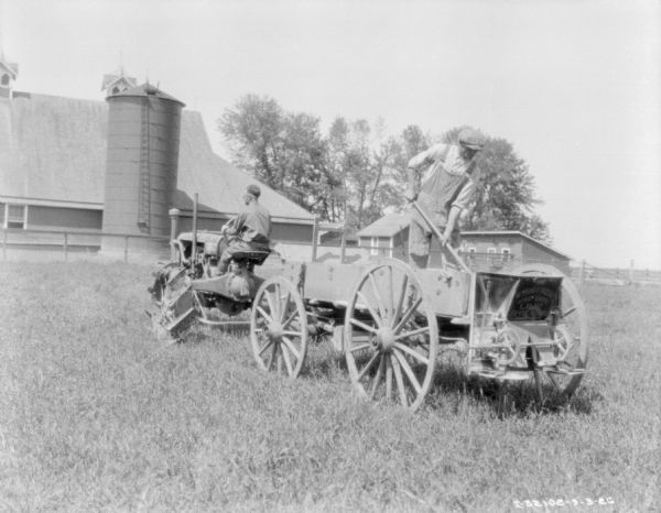 Three-quarter view from left rear of a man driving a tractor pulling a man standing on a lime spreader. In the background is a silo and a barn.