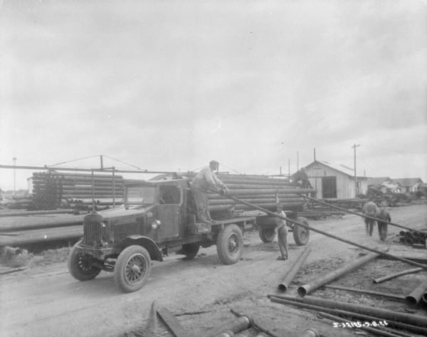 Three men are loading or unloading long pipes with a truck, in a yard with stacks of pipes. Two other men are walking away down the road.