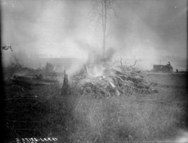 In the foreground is a fire, presumably with stumps that were pulled by the man in the background driving a tractor.