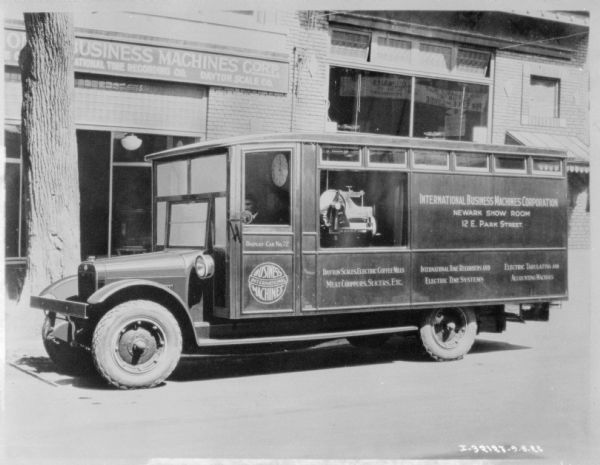 View across street towards an IBM display truck parked along the curb in front of the IBM Business Machine Corp. storefront. A panel behind the driver's side door is open, displaying a machine. A man is sitting in the driver's seat, and there is a large clock behind him in the cab.