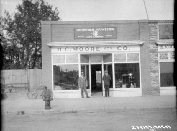View across street towards two men wearing suits and hats standing on the sidewalk in front of a dealership. The sign above the entrance reads: "H.C. Moore and Co."