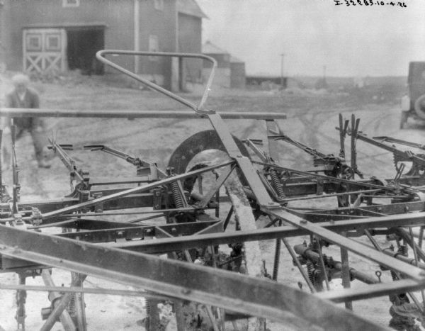 Close-up of a cultivator. In the background is a man standing in a barnyard, and an automobile parked on the far right. In the far background are farm buildings.