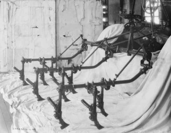 Cultivator set up indoors on a sheet covering the floor beneath it. The sheet is also covering the tractor it is attached to. A white wall and part of the inside of a factory building is in the background.