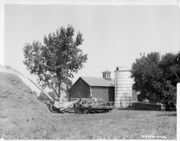 View across field towards a harvester thresher throwing grain on a tall pile on the left. Men are working on the back of a horse-drawn wagon. Farm buildings are in the background.