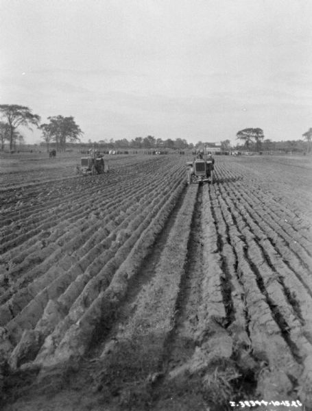 View down freshly plowed field towards men on tractors. There is a crowd of men in the far distance.