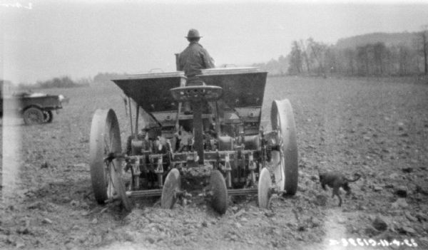 Rear view of a man driving a tractor pulling a potato planter in a field. There is a wagon or truck parked in the background on the left. A dog in the field on the right, running alongside the planter.