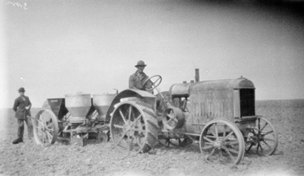 Three-quarter view from front right of a man driving a tractor pulling a potato planter in a field. Another man is standing on the left behind the potato planter.