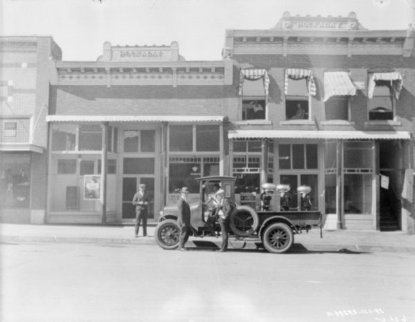 View across street towards three men standing near a man sitting in the driver's seat of a delivery truck parked at the curb. There are storefronts along the sidewalk. Cream separators are loaded in the bed of the truck.