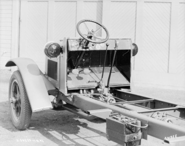 Three-quarter view from left rear of front section of truck with exposed chassis parked in front of a garage with closed doors.