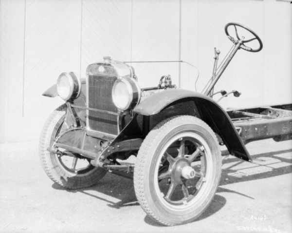 Three-quarter view from front left of the front section of a truck chassis. There is a front grill, headlights, and fenders, and exposed chassis behind the steering wheel.