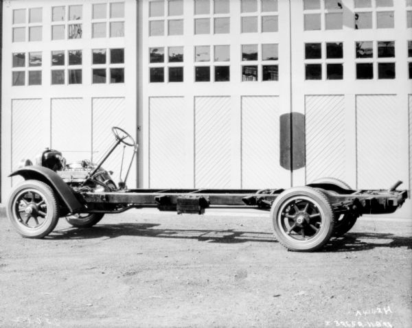 Left side view of a truck with exposed chassis parked in front of a garage with the doors closed.