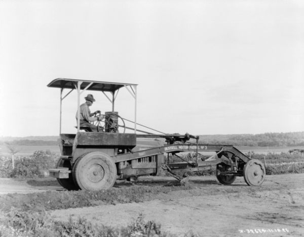 Right side profile view of a man using a grader on an unpaved road.