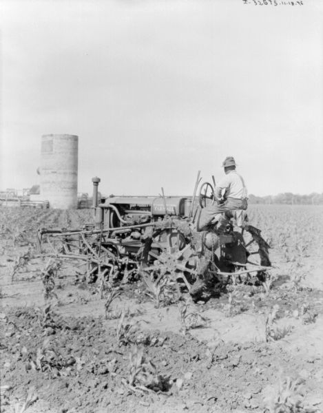 Three-quarter view from left rear of a man driving a Farmall tractor with a mounted cultivator in a field. In the background are farm buildings, and a silo without a roof.