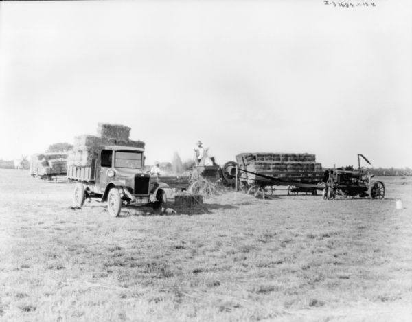 View across field towards a truck, wagons, and a tractor belt-driving a hay press. Bales of hay are stacked on the wagons and the truck. Two men are working near the hay press.