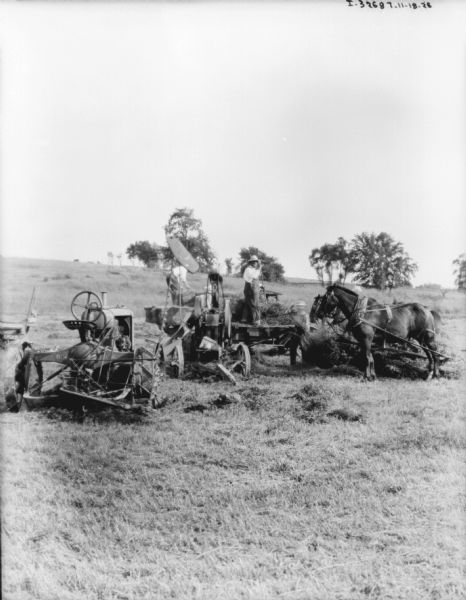 View across field towards a wagon, and a tractor belt-driving a hay press. Three men are working near the hay press.