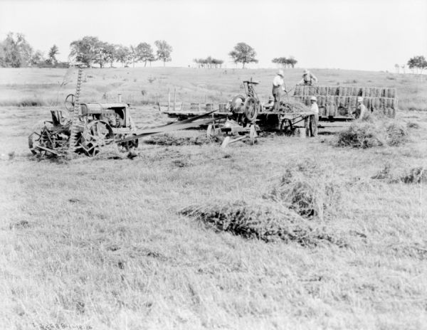 View across field towards a truck, wagons, and a tractor belt-driving a hay press. Bales of hay are stacked on the wagons and the truck. Three men are working near the hay press, another man is standing near a pile of hay, and another man is standing on a wagon stacked with hay bales.