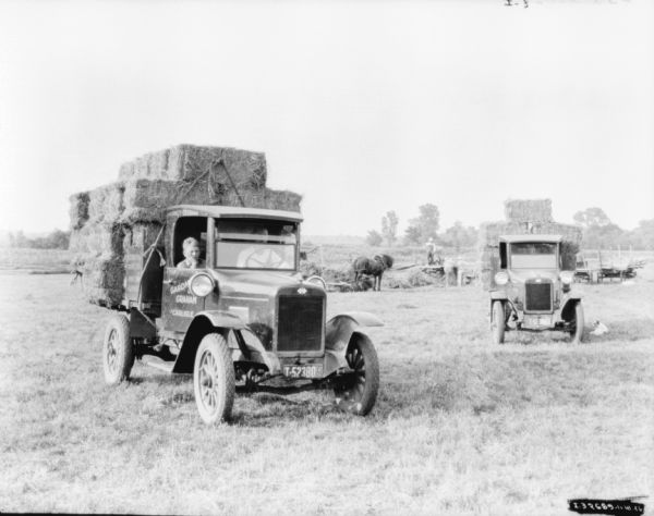 In the foreground on the left is a truck, with a man in the driver's seat, and a boy in the passenger seat. A sign painted on the door of the truck reads: "Gardm... & Graham, Carlisle." There is another truck on the right. In the background in the field are three men standing near a hay press. A horse is standing nearby.