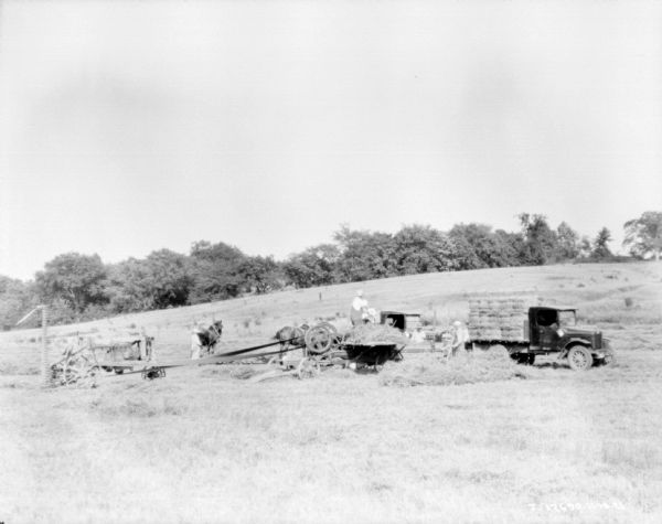 View across field towards men working at a baling operation. On the right is a truck stacked with bales of hay. A man is standing near a hay press, which is being belt-drive by a tractor on the left. A man is standing near a horse in the background.
